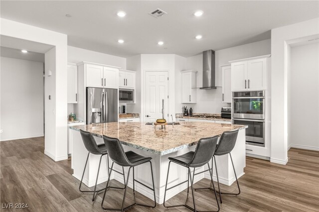 kitchen featuring sink, wall chimney exhaust hood, a kitchen island with sink, white cabinetry, and appliances with stainless steel finishes