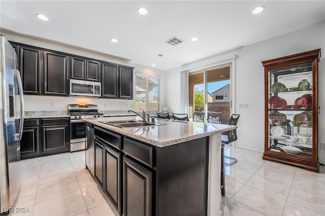 kitchen with appliances with stainless steel finishes, light tile patterned flooring, light stone counters, a center island with sink, and sink