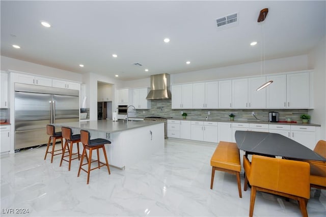 kitchen with high end appliances, wall chimney exhaust hood, white cabinets, and a kitchen island with sink