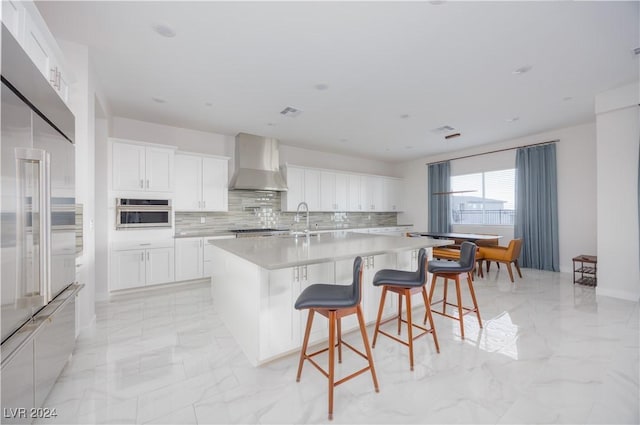 kitchen featuring a breakfast bar, appliances with stainless steel finishes, an island with sink, white cabinets, and wall chimney exhaust hood