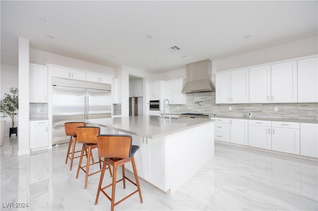 kitchen featuring white cabinetry, stainless steel appliances, a center island with sink, and wall chimney range hood