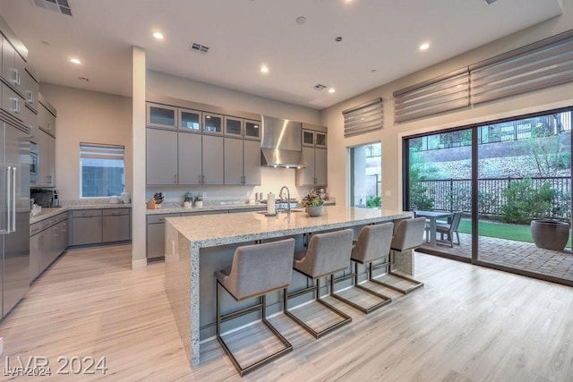 kitchen featuring an island with sink, wall chimney exhaust hood, light wood-type flooring, and gray cabinetry