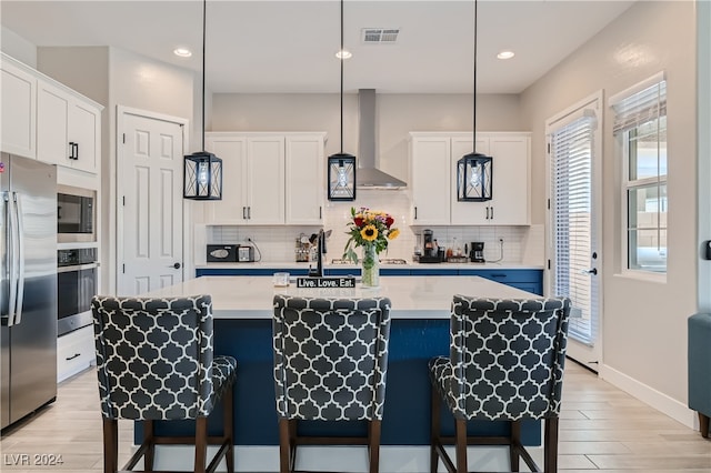 kitchen featuring a kitchen island with sink, wall chimney exhaust hood, stainless steel appliances, and white cabinetry