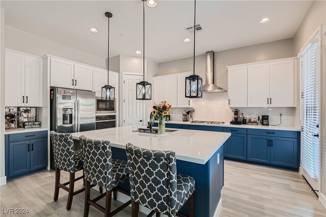 kitchen featuring a kitchen island with sink, wall chimney range hood, appliances with stainless steel finishes, decorative light fixtures, and blue cabinetry