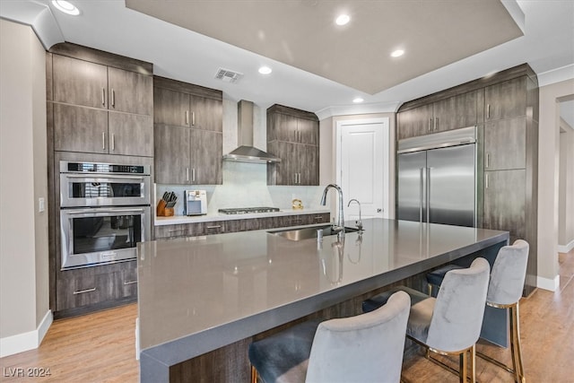 kitchen with a breakfast bar, sink, wall chimney exhaust hood, stainless steel appliances, and a center island with sink