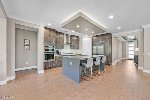 kitchen with a kitchen island with sink, wall chimney exhaust hood, stainless steel appliances, a breakfast bar area, and light hardwood / wood-style floors