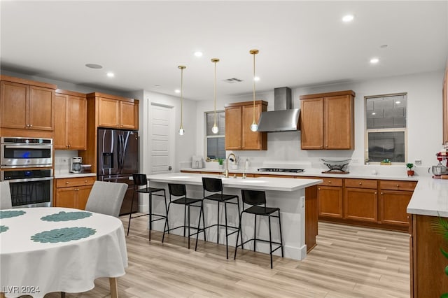 kitchen featuring an island with sink, hanging light fixtures, wall chimney exhaust hood, light hardwood / wood-style flooring, and appliances with stainless steel finishes