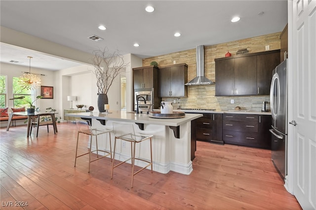 kitchen with an island with sink, dark brown cabinets, wall chimney exhaust hood, light hardwood / wood-style flooring, and appliances with stainless steel finishes