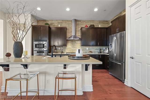 kitchen with a breakfast bar area, wall chimney range hood, appliances with stainless steel finishes, dark brown cabinetry, and dark hardwood / wood-style flooring