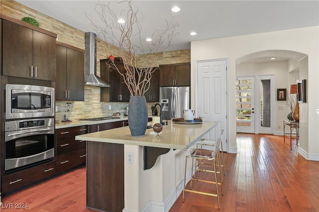 kitchen with an island with sink, wall chimney exhaust hood, a kitchen breakfast bar, appliances with stainless steel finishes, and hardwood / wood-style floors