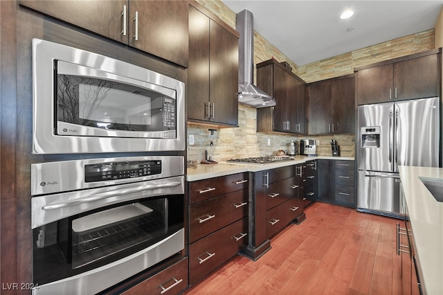 kitchen with tasteful backsplash, dark brown cabinets, wall chimney range hood, appliances with stainless steel finishes, and light hardwood / wood-style floors