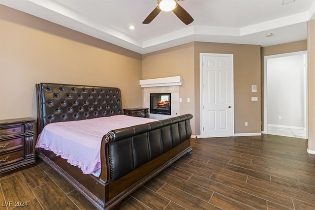 bedroom with a fireplace, a tray ceiling, ceiling fan, and dark wood-type flooring