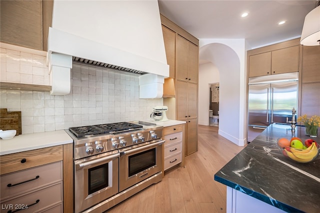 kitchen featuring high quality appliances, backsplash, dark stone countertops, premium range hood, and light hardwood / wood-style floors