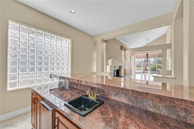 kitchen with dark stone countertops, a multi sided fireplace, vaulted ceiling, decorative light fixtures, and sink