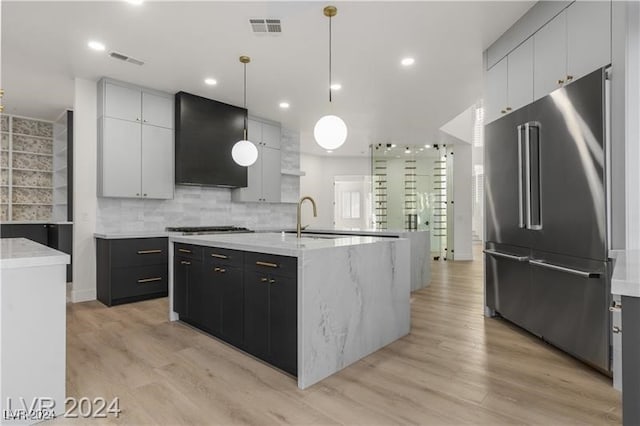 kitchen with pendant lighting, custom exhaust hood, an island with sink, light hardwood / wood-style flooring, and appliances with stainless steel finishes