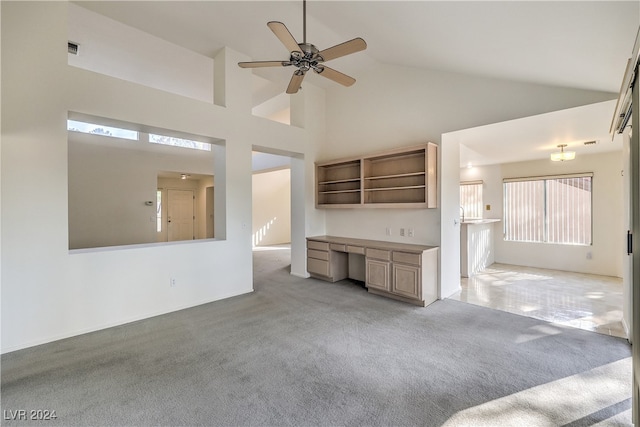 unfurnished living room with ceiling fan, light colored carpet, built in desk, and high vaulted ceiling