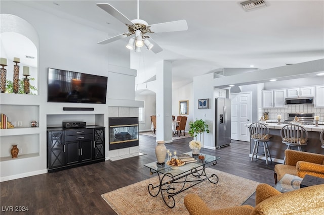 living room with ceiling fan, built in features, a tiled fireplace, dark wood-type flooring, and high vaulted ceiling