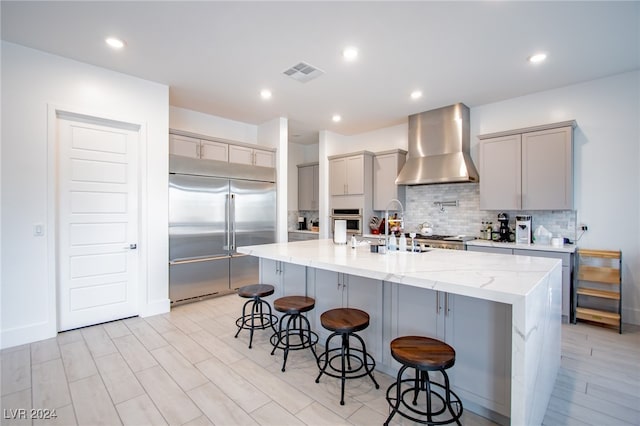 kitchen with appliances with stainless steel finishes, an island with sink, a breakfast bar, backsplash, and wall chimney exhaust hood