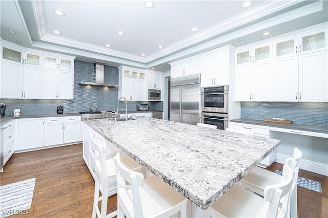 kitchen featuring a breakfast bar, wall chimney range hood, built in appliances, a center island with sink, and white cabinets