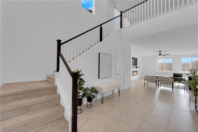 stairway featuring a high ceiling, a ceiling fan, a glass covered fireplace, baseboards, and tile patterned floors