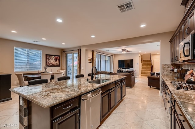 kitchen with light stone counters, sink, a center island with sink, appliances with stainless steel finishes, and dark brown cabinetry