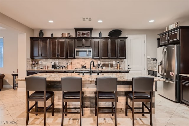 kitchen with dark brown cabinetry, a kitchen island with sink, light stone countertops, and stainless steel appliances