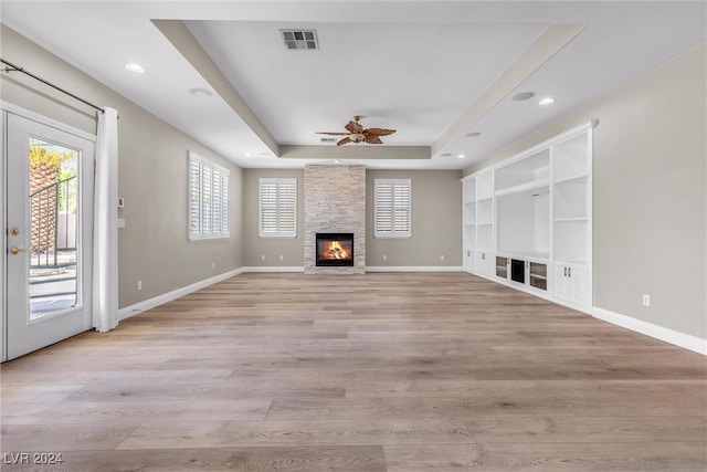 unfurnished living room with ceiling fan, a raised ceiling, a stone fireplace, built in features, and light wood-type flooring