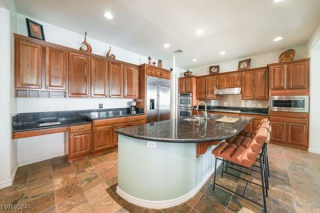 kitchen featuring sink, a center island with sink, built in appliances, a kitchen bar, and decorative backsplash