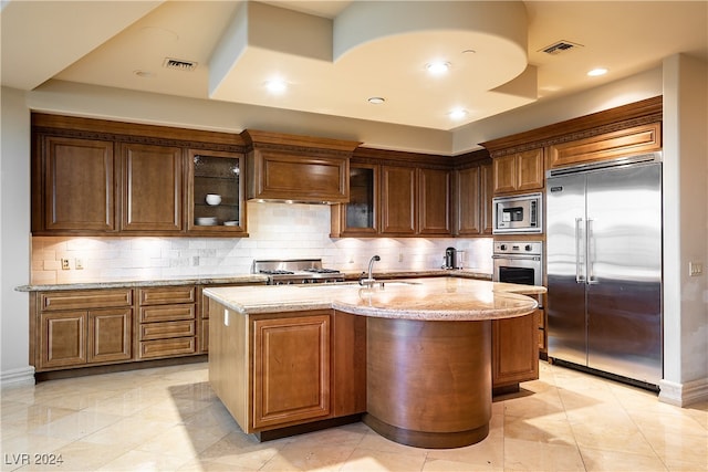 kitchen with light stone countertops, a kitchen island with sink, built in appliances, and tasteful backsplash