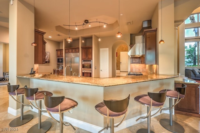 kitchen featuring built in appliances, wall chimney range hood, kitchen peninsula, pendant lighting, and a breakfast bar area