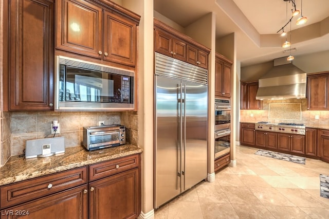 kitchen with backsplash, built in appliances, wall chimney range hood, and light stone counters