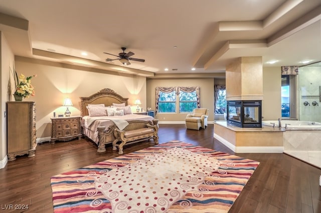 bedroom with ceiling fan, a multi sided fireplace, a tray ceiling, and dark hardwood / wood-style flooring