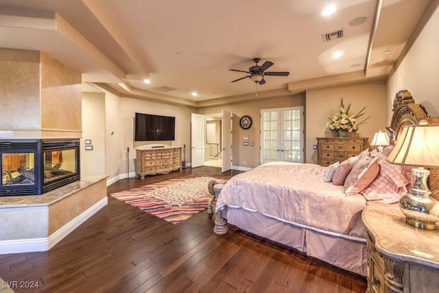 bedroom featuring ceiling fan, a multi sided fireplace, dark hardwood / wood-style flooring, and a raised ceiling