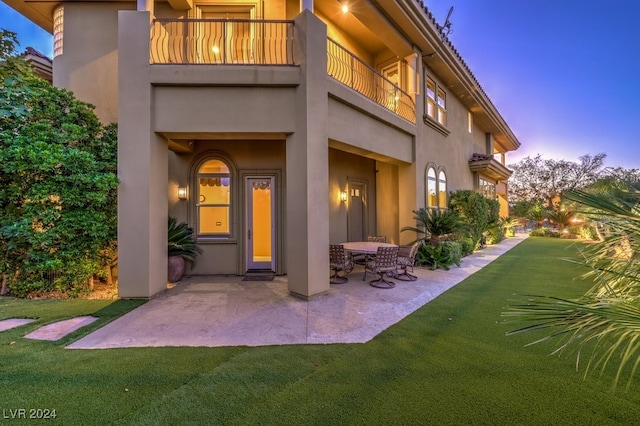 back house at dusk featuring a balcony, a patio, and a lawn