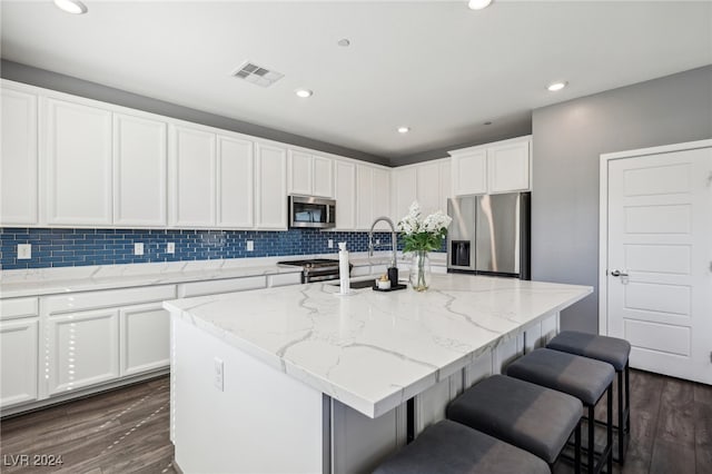 kitchen with appliances with stainless steel finishes, a center island with sink, dark hardwood / wood-style flooring, and light stone countertops