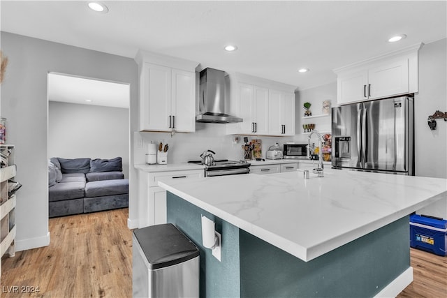 kitchen with white cabinets, wall chimney exhaust hood, an island with sink, light wood-type flooring, and appliances with stainless steel finishes