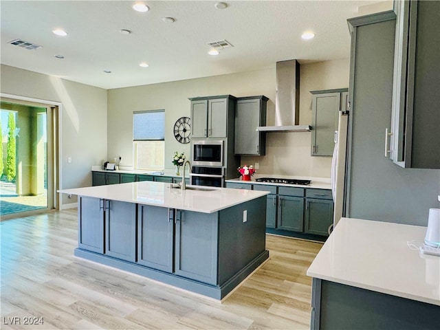 kitchen with light hardwood / wood-style floors, an island with sink, wall chimney exhaust hood, stainless steel appliances, and sink