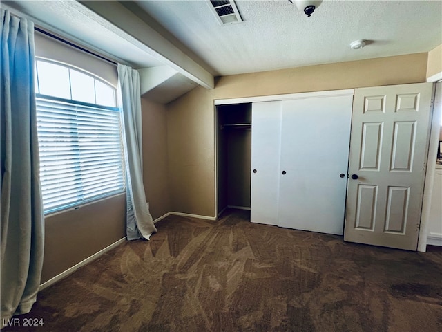 unfurnished bedroom with a textured ceiling, a closet, lofted ceiling with beams, and dark colored carpet