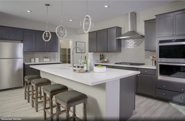 kitchen featuring appliances with stainless steel finishes, an island with sink, backsplash, wall chimney exhaust hood, and light wood-type flooring