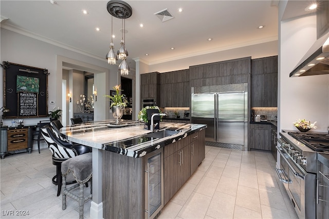 kitchen with ornamental molding, wall chimney exhaust hood, high end appliances, a large island with sink, and a breakfast bar area