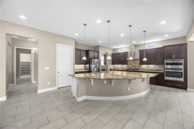 kitchen with pendant lighting, a spacious island, wall chimney range hood, appliances with stainless steel finishes, and a kitchen bar