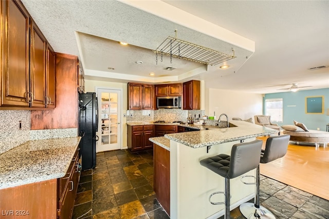 kitchen featuring light stone counters, stainless steel appliances, kitchen peninsula, a tray ceiling, and a breakfast bar area