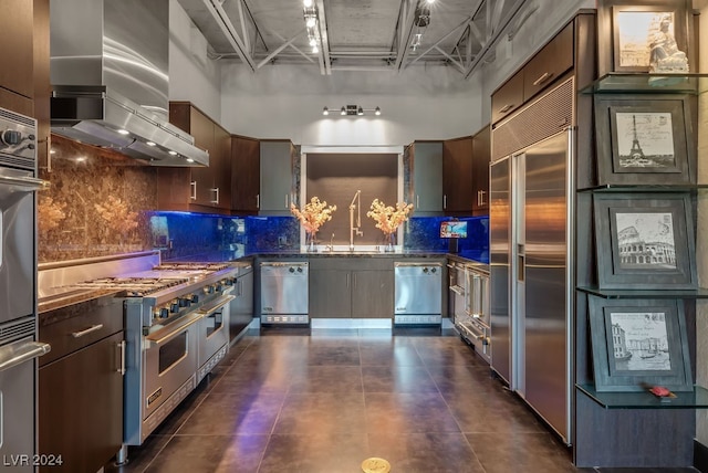 kitchen with sink, dark brown cabinets, premium appliances, backsplash, and wall chimney range hood