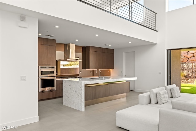 kitchen featuring a kitchen island with sink, wall chimney range hood, sink, appliances with stainless steel finishes, and a towering ceiling