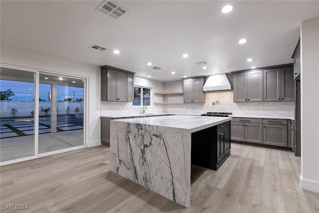 kitchen with a kitchen island, light stone countertops, light wood-type flooring, premium range hood, and backsplash