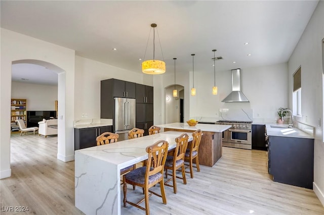 kitchen featuring pendant lighting, high end appliances, wall chimney exhaust hood, light stone countertops, and light wood-type flooring