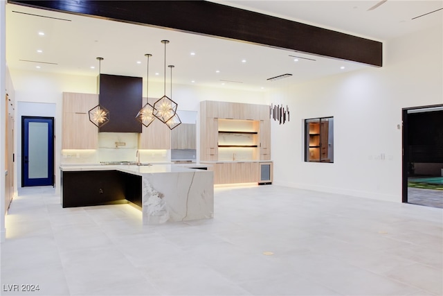 kitchen with custom range hood, light brown cabinets, pendant lighting, light stone countertops, and a center island with sink