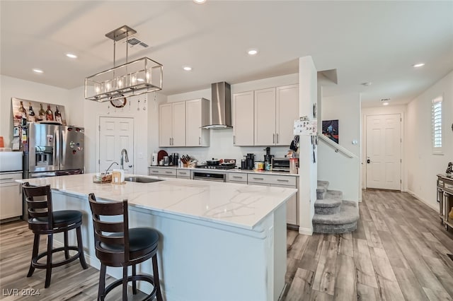 kitchen with appliances with stainless steel finishes, light hardwood / wood-style flooring, a center island with sink, sink, and wall chimney range hood