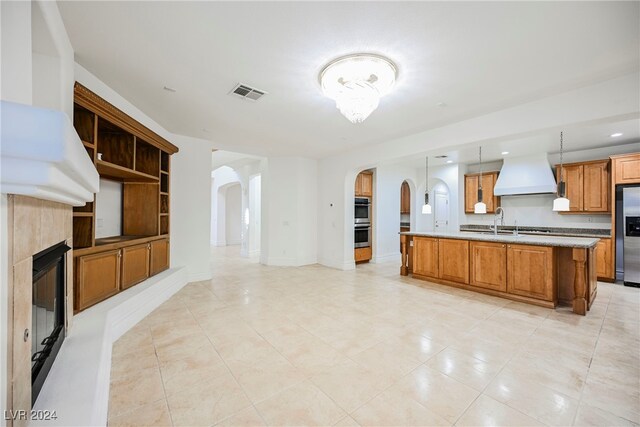 kitchen with a center island with sink, appliances with stainless steel finishes, custom range hood, a tile fireplace, and pendant lighting