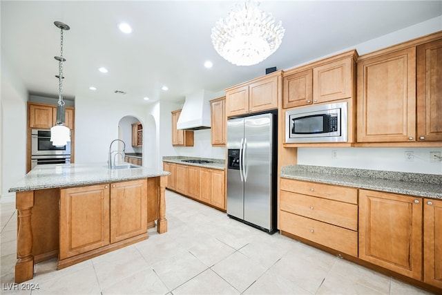 kitchen featuring appliances with stainless steel finishes, hanging light fixtures, light stone counters, premium range hood, and a center island with sink